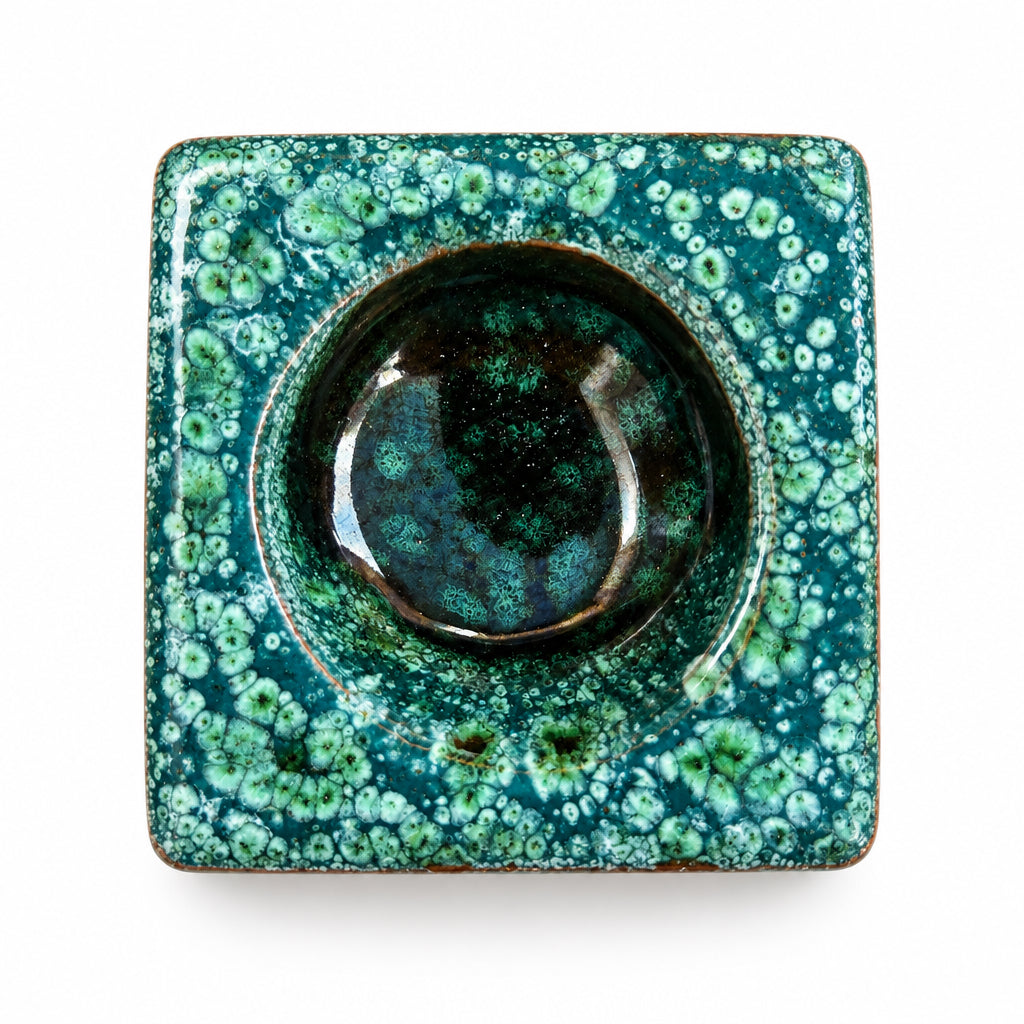 Brûle-parfum carré en céramique vert émeraude et marron, avec finition mouchetée, motifs ajourés en losanges et bougie chauffe-plat allumée à l’intérieur.
