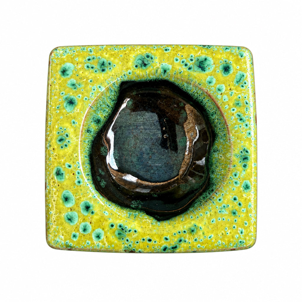 Brûle-parfum carré en céramique verte et marron, avec finition mouchetée, motifs ajourés et bougie chauffe-plat allumée.