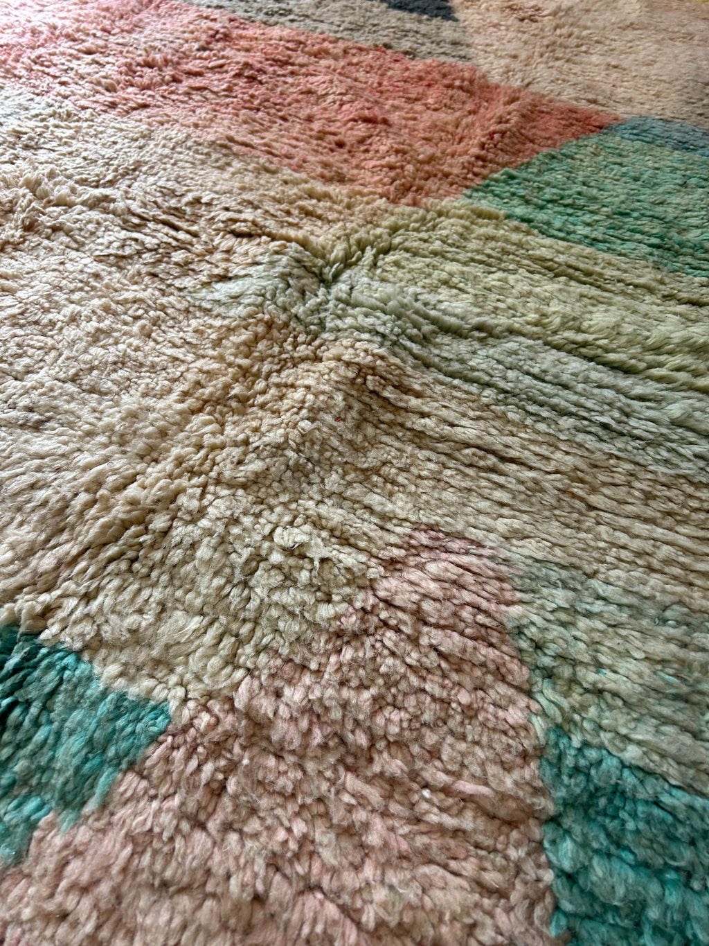 Tapis Beni Ourain fait main dans un salon contemporain – motifs géométriques pastel rose saumon, bleu vert et ivoire avec franges, table basse dorée en arrière-plan