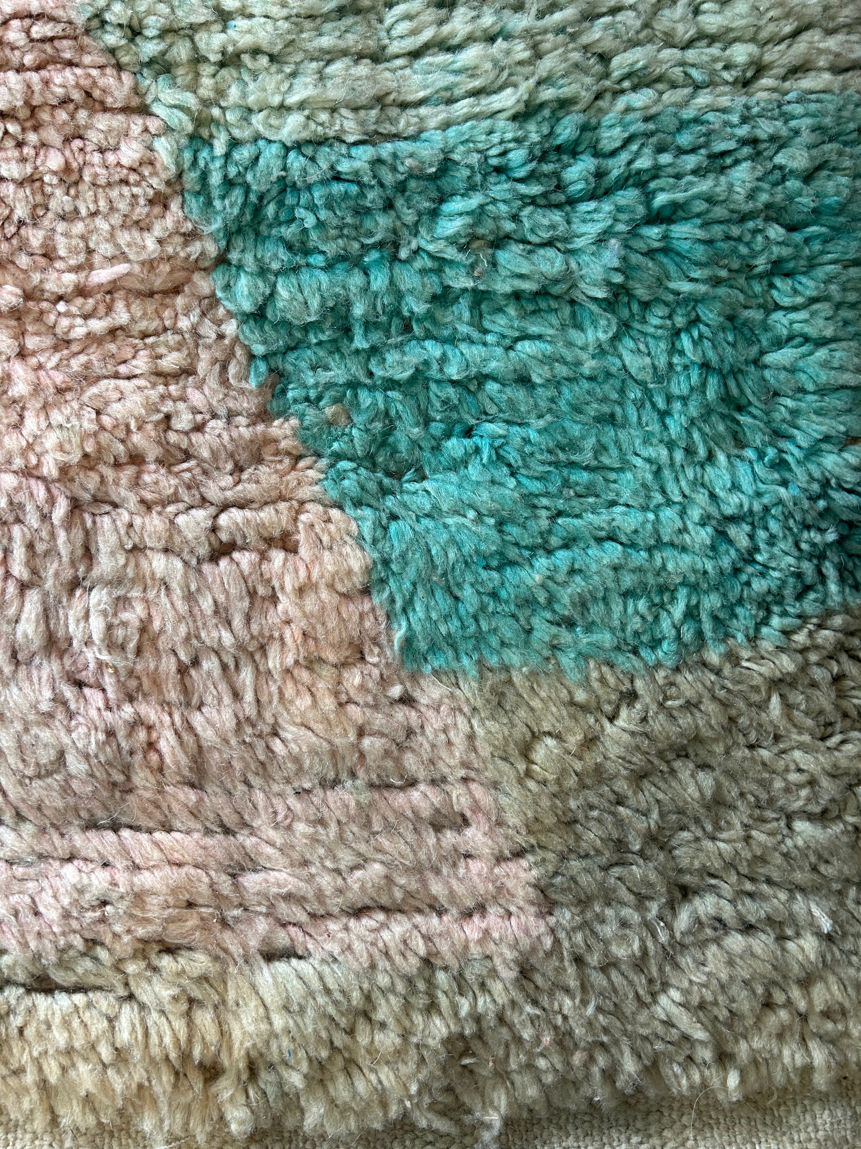 Tapis Beni Ourain marocain posé sur une terrasse en terre battue – motifs géométriques pastels rose, bleu vert et triangle anthracite, palmiers et Atlas enneigé en arrière-plan