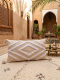 Housse de coussin XL lombaire beige sable, motifs losanges tufted blanc et pompons, posée dans un riad marocain – décoration boho-luxe fait main