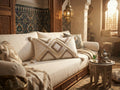 "Housse de coussin berbère XL blanc écru et sable avec pompons posée sur un canapé blanc dans un salon marocain luxe, zellige et bois sculpté en décor"