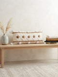 Housse de coussin XL berbère 98x48 cm, motif losanges et médaillons camel sur fond ivoire, pompons blancs, posée sur coffre sculpté dans un riad marocain