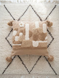  Housse de coussin berbère punch needle abstraite 48x48 cm, tons sable et ivoire, pompons camel, posée sur un tapis dans un riad marocain