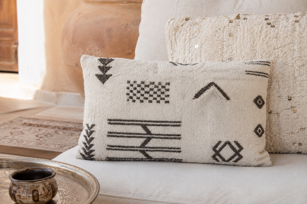 Housse de coussin berbère ivoire sur banquette riad – motifs noirs traditionnels au soleil du Maroc