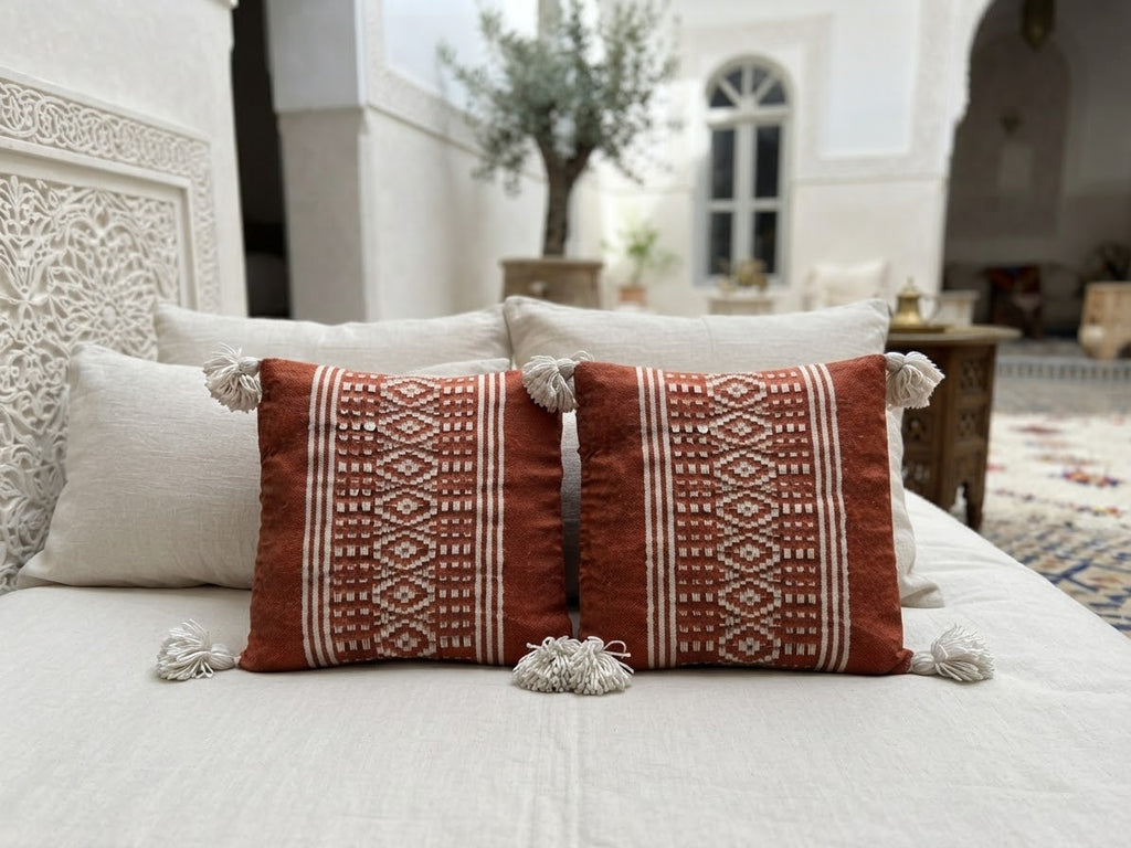 Duo de housses de coussin marocaines terracotta et écru sur lit blanc – Tissage berbère fait main avec pompons